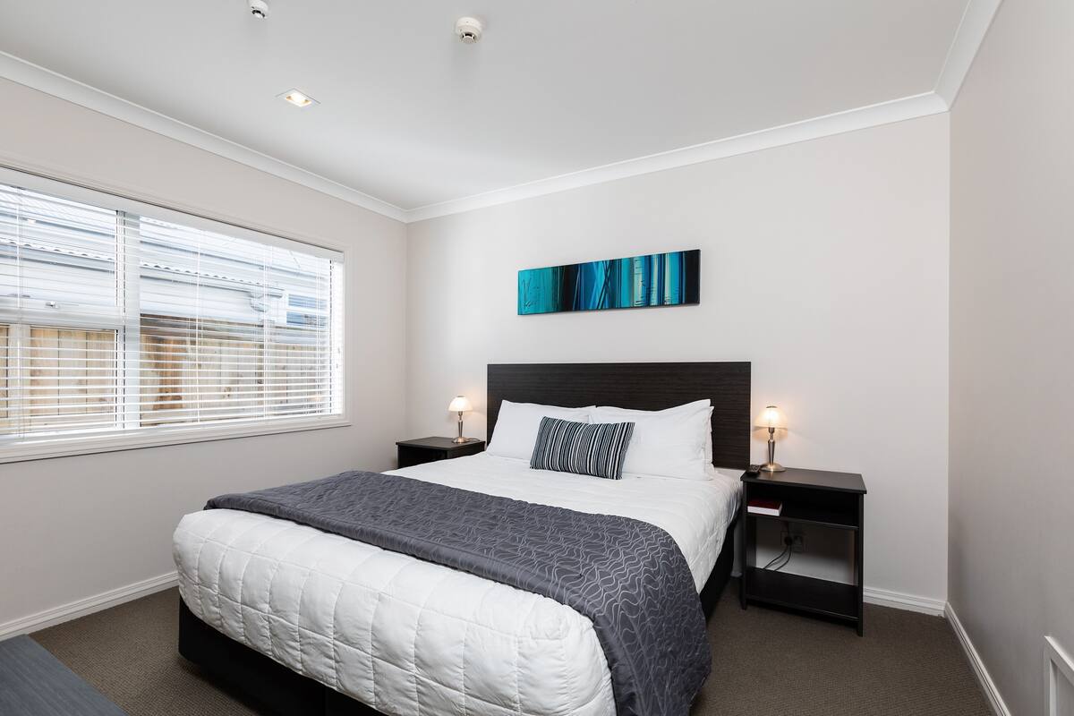 A bedroom with a bed, two bedside tables, a nightstand, a window with blinds, and a wall-mounted artwork.