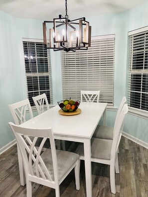 Un coin repas avec une table blanche, des chaises blanches, un panier de fruits et un lustre moderne.