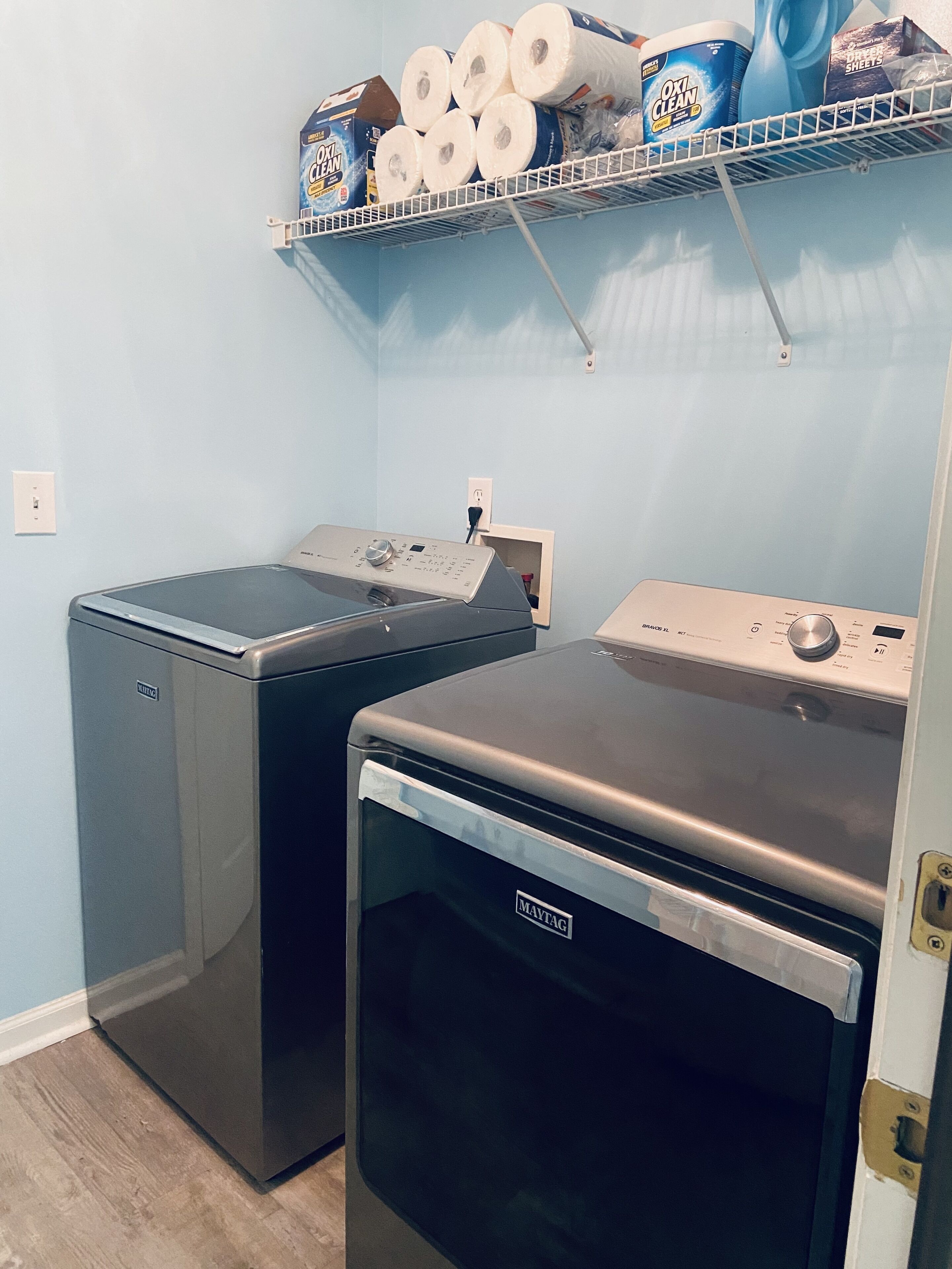 A laundry room with two Maytag washers and dryers, a shelf with laundry supplies, and a power outlet.