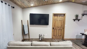 Um quarto com uma televisão de tela grande fixada na parede, uma porta de madeira, um sofá e uma pequena mesa com uma planta decorativa.