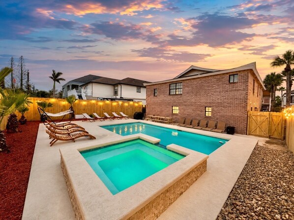A rectangular swimming pool with a small hot tub, surrounded by lounge chairs and a wooden deck.