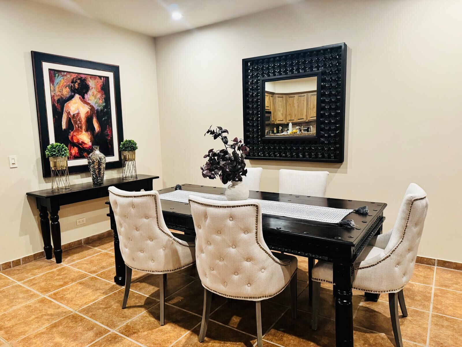 Zona pranzo con tavolo nero, quattro sedie imbottite, un vaso con fiori, un quadro incorniciato e uno specchio decorativo.