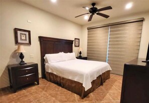 Quarto com cama, mesas de cabeceira, ventoinha no teto e uma janela grande com persianas.