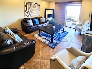 Sala de estar com um sofá de couro preto, uma mesa de centro e uma poltrona bege. Há uma janela grande com vista para o exterior.