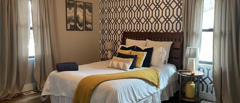 Un dormitorio con suelo de madera, cama con ropa de cama blanca y varios cojines, ventilador de techo y pared estampada.