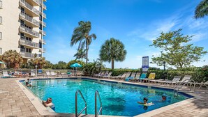 A swimming pool with lounge chairs and umbrellas, surrounded by palm trees and a tall building. - Direct beachfront! Best gulf views and sunsets! Pickleball and tennis!  (Fort Myers Beach)