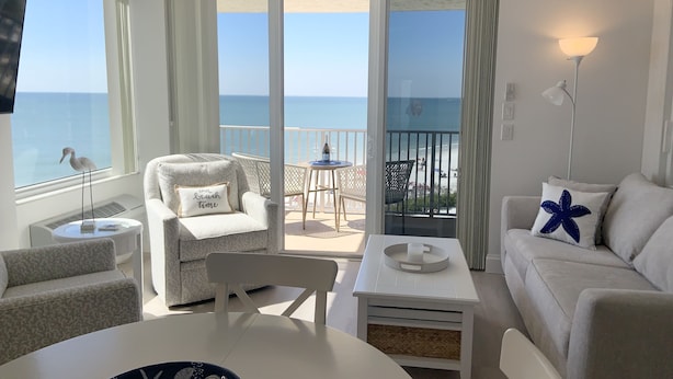 A modern living room with a view of the sea, a balcony with a table and chairs, and a ceiling fan. - Direct beachfront! Best gulf views and sunsets! Pickleball and tennis!  (Fort Myers Beach)