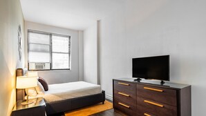 A bedroom with a bed, a TV on a dresser, a window with blinds, and an air conditioning unit. - Timeless NYC Living | UWS | Pet-Friendly (New York)