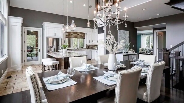 A spacious dining area with a large table set for six, surrounded by high-backed chairs. A chandelier hangs above, and the room features a staircase leading to an upper level. - The Mansion, Wasaga beach, Blue mountain (Utopia)