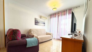 A living room with a purple sofa, a beige sofa, a wooden side table with a TV, and a window with striped curtains.