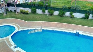 A large, clear swimming pool with a curved design, surrounded by a low brick wall and a green fence.