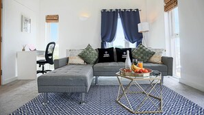 A modern living room with a grey sectional sofa, a matching ottoman, and a coffee table with fruit. There are two black pillows with "YOKO PROPERTY" branding, a wicker basket, and a lamp on a side table. - Duplex Apartment • Balcony • FreeParking • SmartTV (Newport Pagnell)
