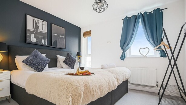 A modern bedroom with a large bed, dark walls, and a window with blue curtains. - Duplex Apartment • Balcony • FreeParking • SmartTV (Newport Pagnell)