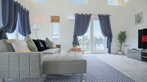 A modern living room with a grey sofa, a coffee table, and a TV. There are large windows with blue curtains, a lamp, and a potted plant. - Duplex Apartment • Balcony • FreeParking • SmartTV (Newport Pagnell)