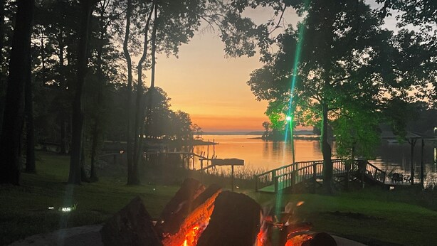 A campfire in a circular stone fire pit surrounded by a grassy area with trees and a body of water in the background. - Quiet Cove with Big Water Views (Ridgeway)