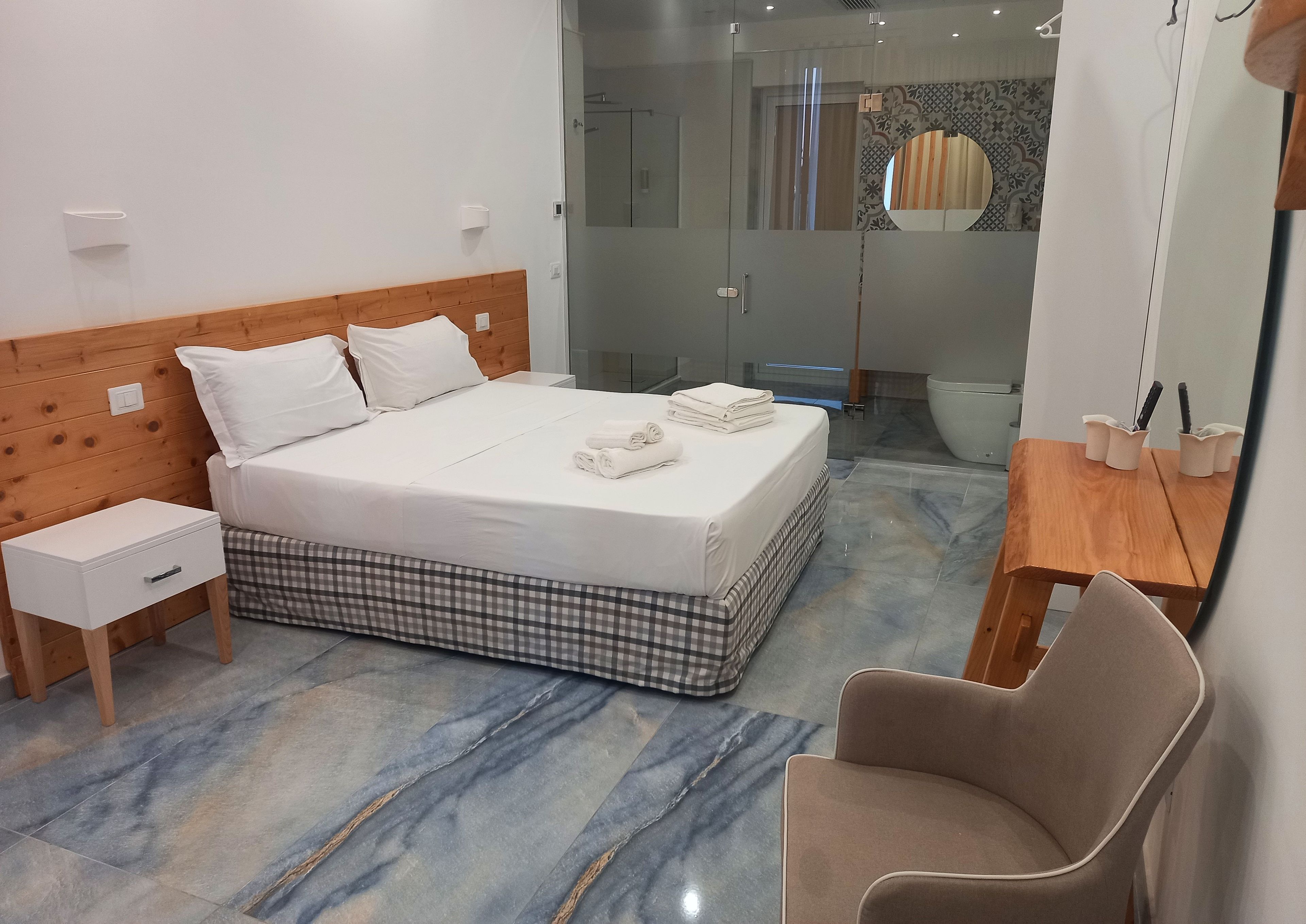 Une chambre d’hôtel moderne avec une tête de lit en bois, un lit recouvert de draps blancs, un couvre-lit à carreaux, une table de chevet, une chaise, un bureau en bois avec un miroir et une salle de bain dotée d’une cabine de douche en verre.