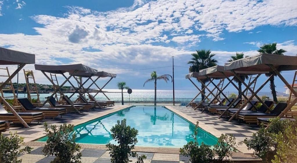 Zona de piscina con sillas de descanso y sombrillas, cielo despejado y vista al mar.
