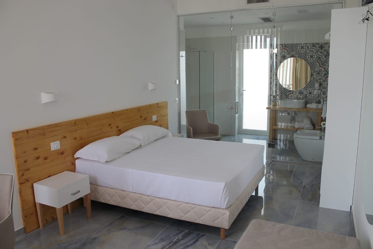 Un dormitorio moderno con cabecera de madera, una cama individual, una mesita de noche y un baño visible a través de una puerta abierta.