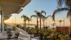 A modern outdoor seating area with white chairs and tables, surrounded by lush greenery and palm trees, overlooking a body of water. - Jonium Hotel (Pulsano)