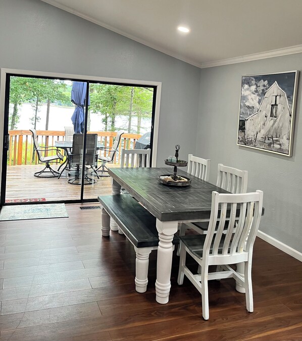 A dining area with a dark wooden table and white chairs, a painting on the wall, and a view through a glass door to an outdoor deck.