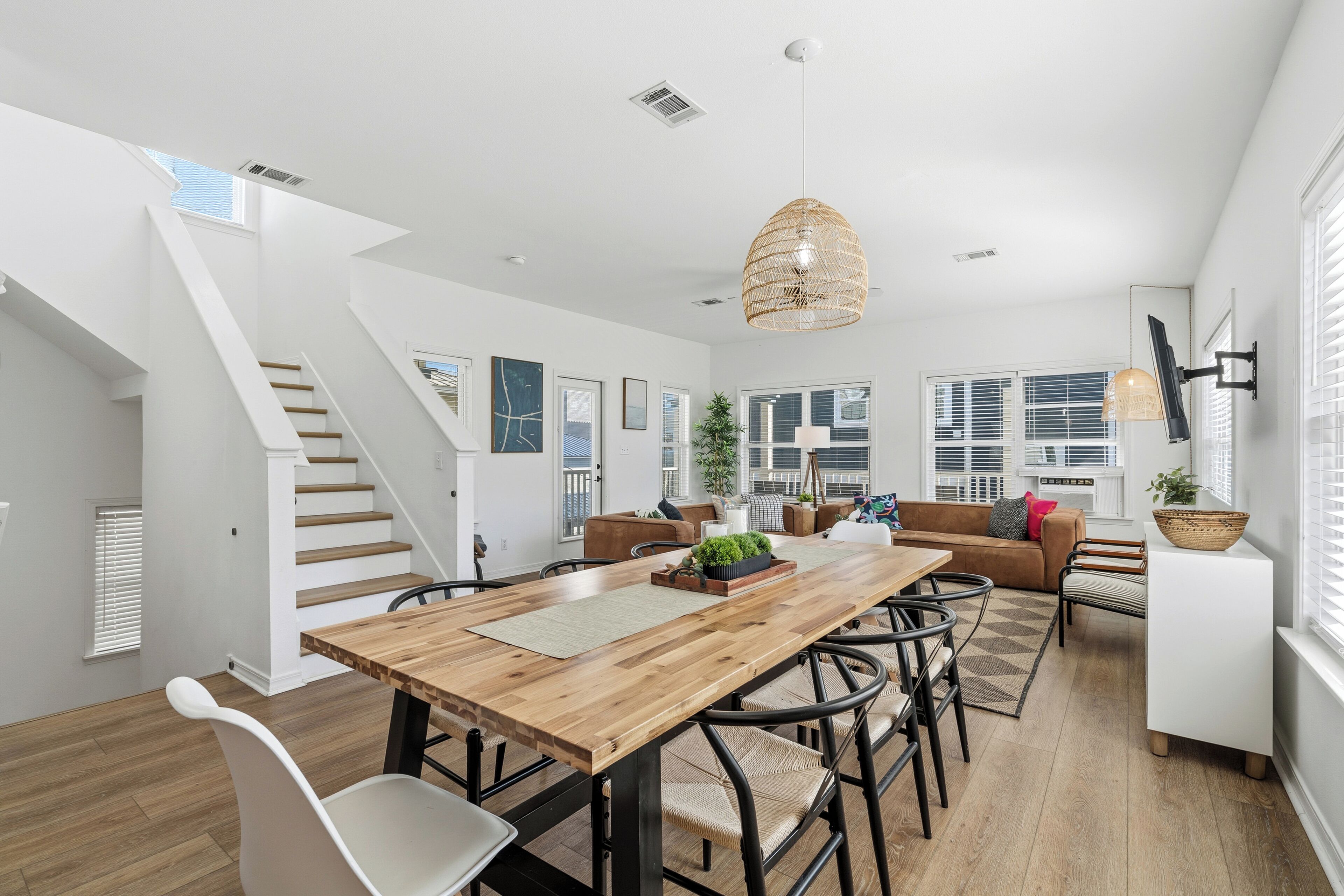 A spacious living area with a wooden dining table, modern chairs, a staircase, and a large window with blinds.