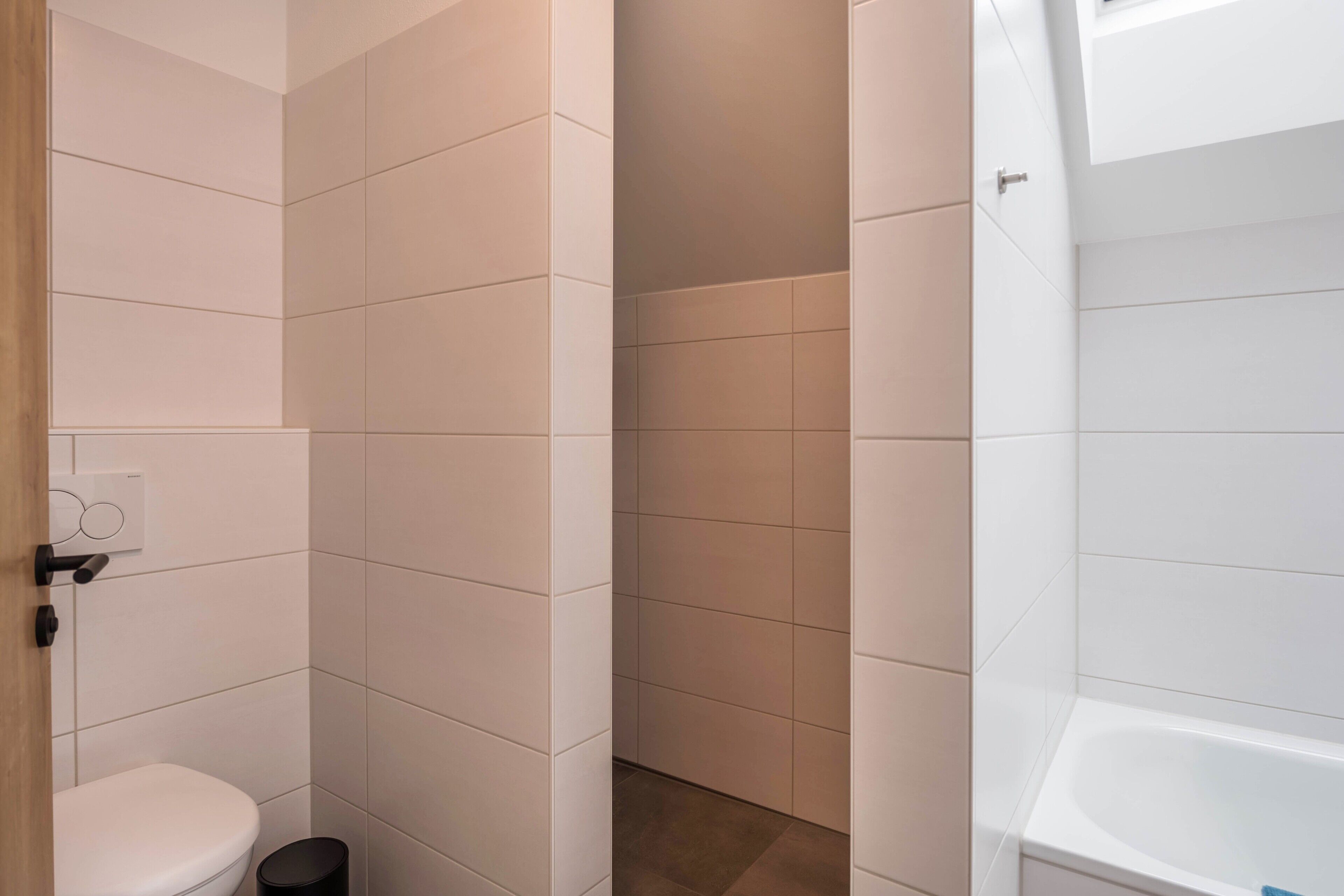 A bathroom with white tiles, a toilet, a trash bin, and a bathtub.