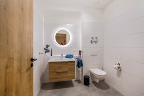Un bagno con porta in legno, uno specchio rotondo, un lavandino, un water e un cestino.