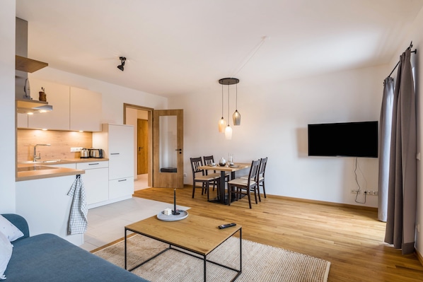 A modern apartment with a kitchen, dining area, and living space, featuring a flat-screen TV, wooden flooring, and a minimalist design.