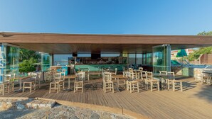 An outdoor dining area with wooden tables and chairs, a bar in the background, and a stone wall in the foreground. - Iolida Village Hotel (Chania)