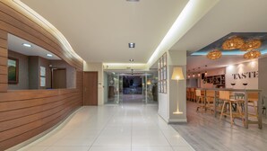 A modern hotel lobby with a wooden reception desk, a glass partition, and a bar area with wooden chairs and a 'TASTE' sign. - Iolida Village Hotel (Chania)