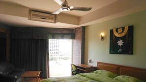 Quarto de hotel com cama, escrivaninha, cadeira, ventilador de teto e janela com cortinas.