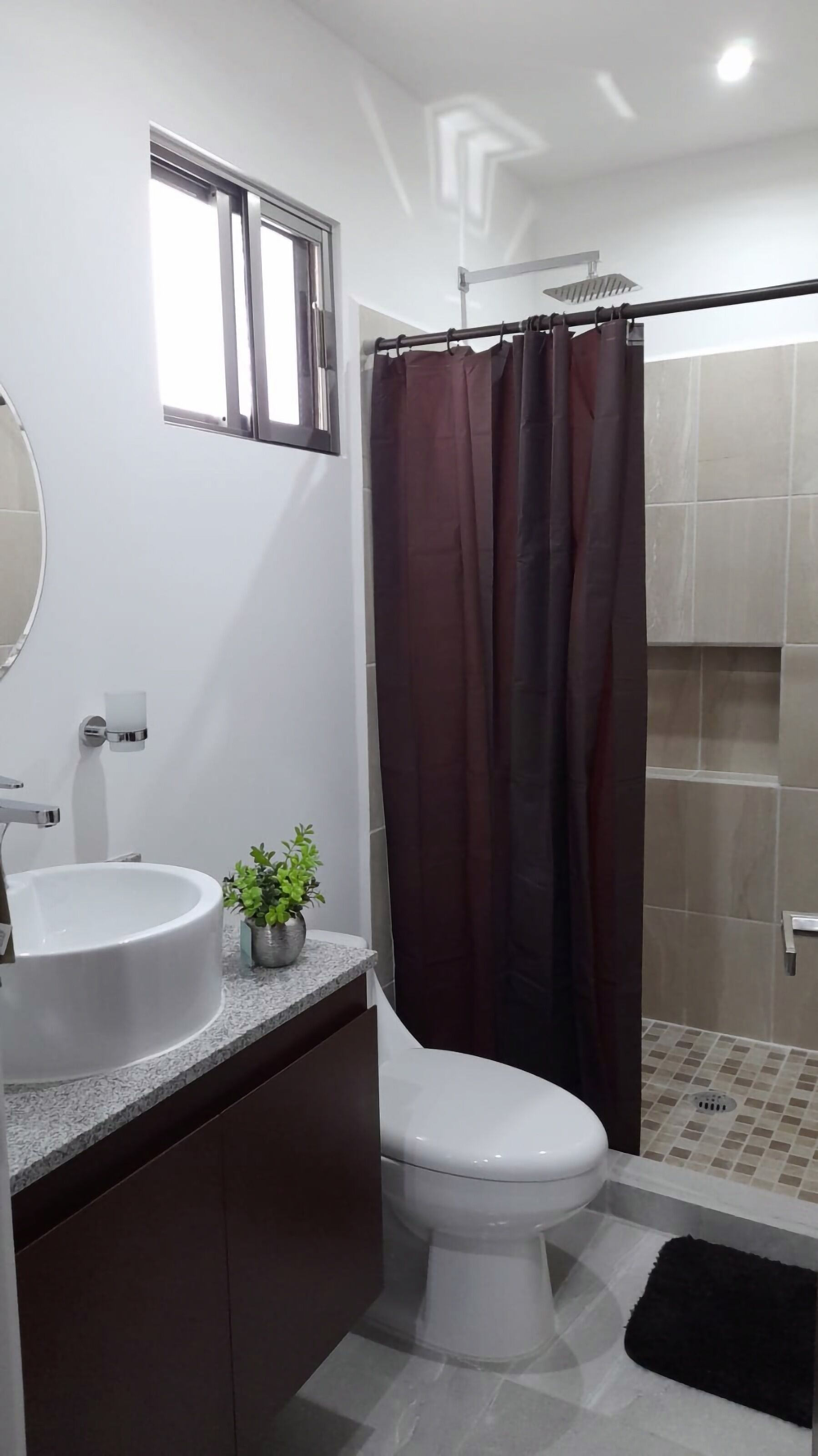 A modern bathroom with a white toilet, a sink with a granite countertop, a small potted plant, a shower curtain, and a tiled shower area.