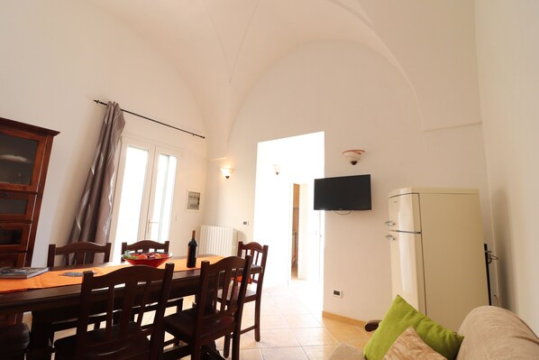 A dining area with a table, chairs, a bowl of fruit, a bottle of wine, and a television mounted on the wall.