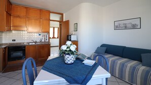 A compact kitchen with wooden cabinets, a microwave, and a sink. There's a dining table with a blue tablecloth and a vase of flowers. A striped sofa is against the wall with a framed picture above it. - Casa Sophia Panoramic Terrace and Elegant Stay in the Heart of Otranto (Otranto)