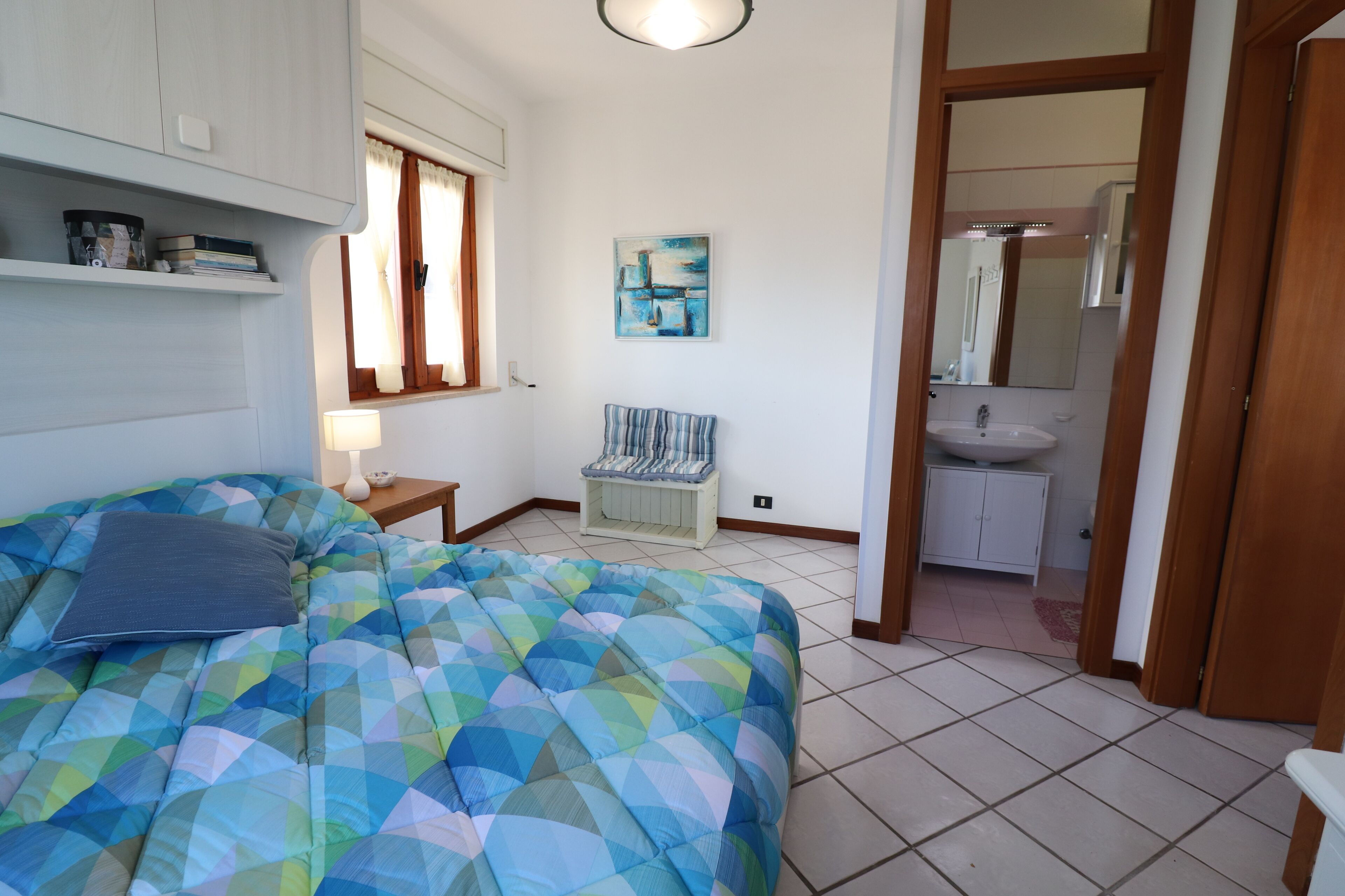 Un dormitorio con cama, mesita de noche, ventana con cortinas, un cuadro colgado en la pared y un baño visible a través de una puerta abierta.