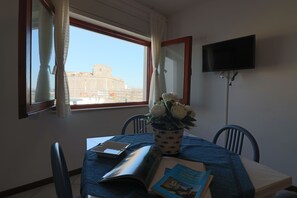 Quarto com mesa de jantar, cadeiras, planta em vaso, um livro, controle remoto e TV na parede.
