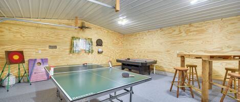 Een tafeltennistafel met krukken, een dartbord en nog een dartbord in een kamer met houten wanden en een metalen plafond.