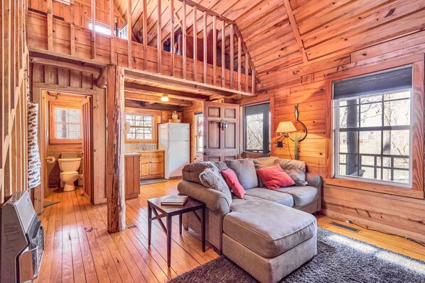 A wooden cabin interior with a loft bedroom, a living room with a sofa and ottoman, and a kitchen area.