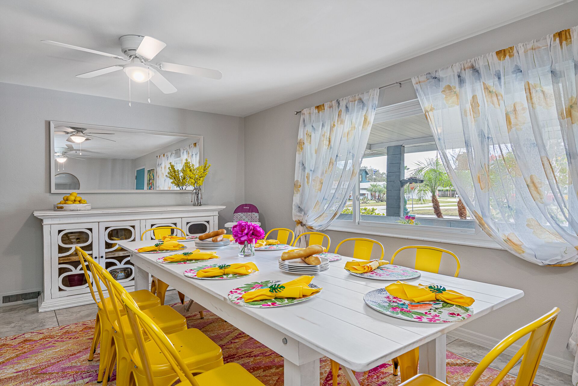 A dining area with a white table set for four, yellow chairs, a ceiling fan, and a window with floral curtains.