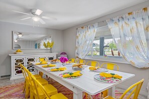Comedor con una mesa blanca dispuesta para cuatro, sillas amarillas, un ventilador de techo y una ventana con cortinas florales.