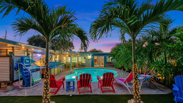 A backyard with a swimming pool, red Adirondack chairs, and palm trees.