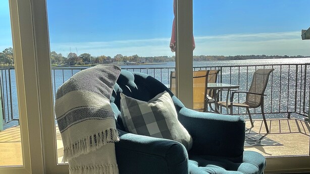 A cozy seating area with a dark blue sofa, a patterned throw blanket, and a view of a lake through large glass doors. - New! Lakefront 2-bedroom Condo. Newly remodeled. (Willis)