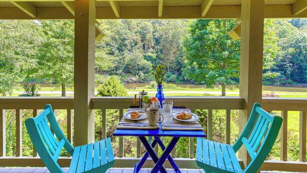 Uma área de estar coberta ao ar livre, com uma mesa posta para dois, oferecendo vista para um jardim exuberante e um corpo d'água.