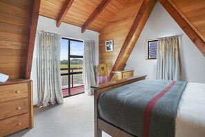 A bedroom with a wooden ceiling, a bed with a grey blanket, a wooden dresser, and a view of a green landscape through a large window. - Snow Chalet - Fairlie Holiday Home (Fairlie)