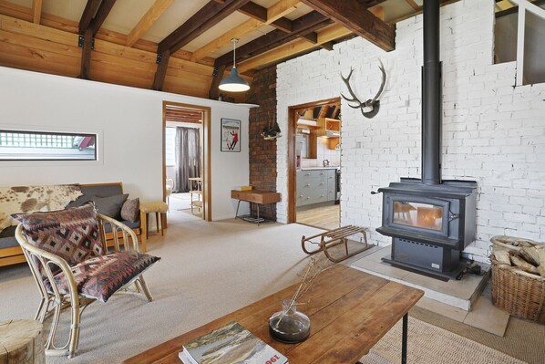 A cozy living room with a fireplace, a sofa, a coffee table, and a dining area with a brick wall and wooden beams. - Snow Chalet - Fairlie Holiday Home (Fairlie)