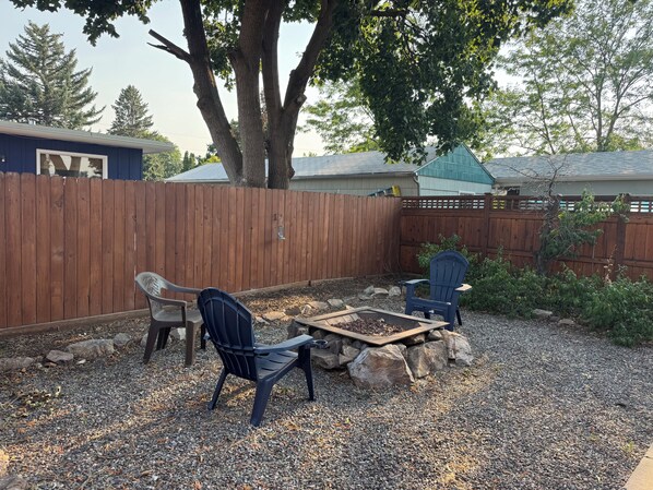 A backyard with a wooden fence, two Adirondack chairs, a fire pit, and a gravel ground.