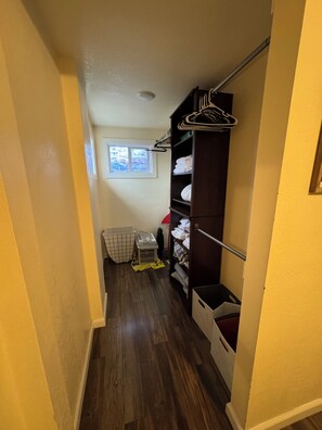 A narrow hallway with wooden flooring, a closet with hanging clothes, a laundry basket, and a window with blinds.