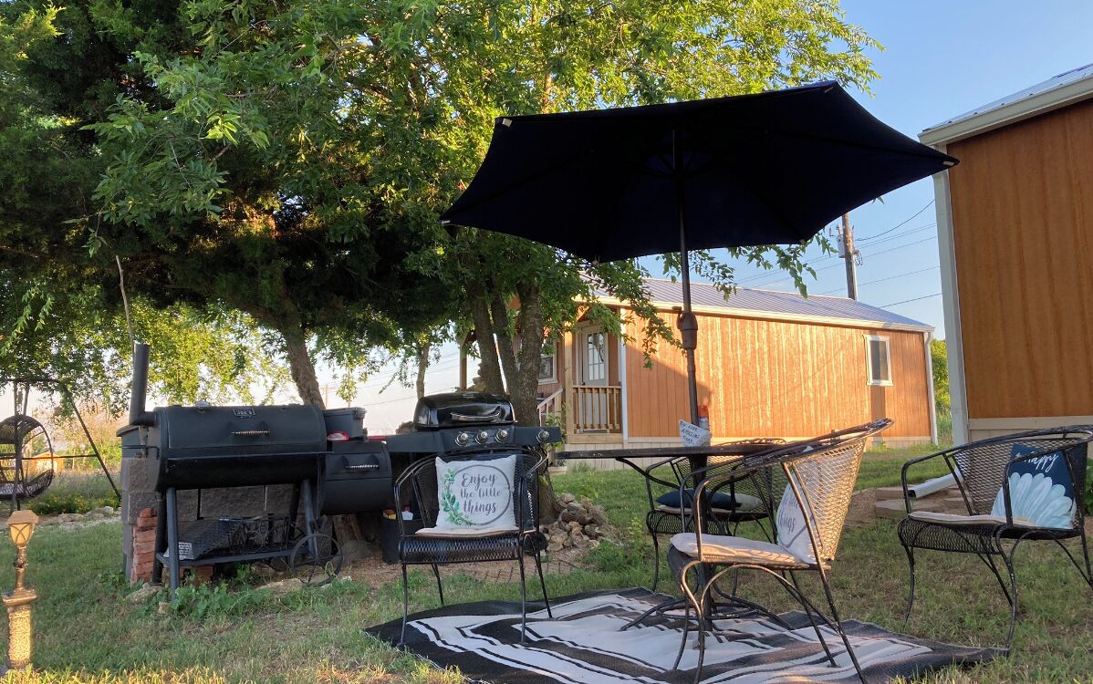 En udendørs terrasse med grill, stole og bord under en stor parasol.