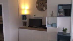 En moderne stue med et fladskærms-tv, træskabe, et ur og reoler med bøger og dekorative genstande.
