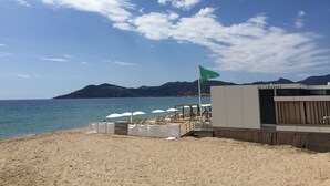 En bygning ved stranden med et flag, strandstole, parasoller og udsigt til havet og bjergene.